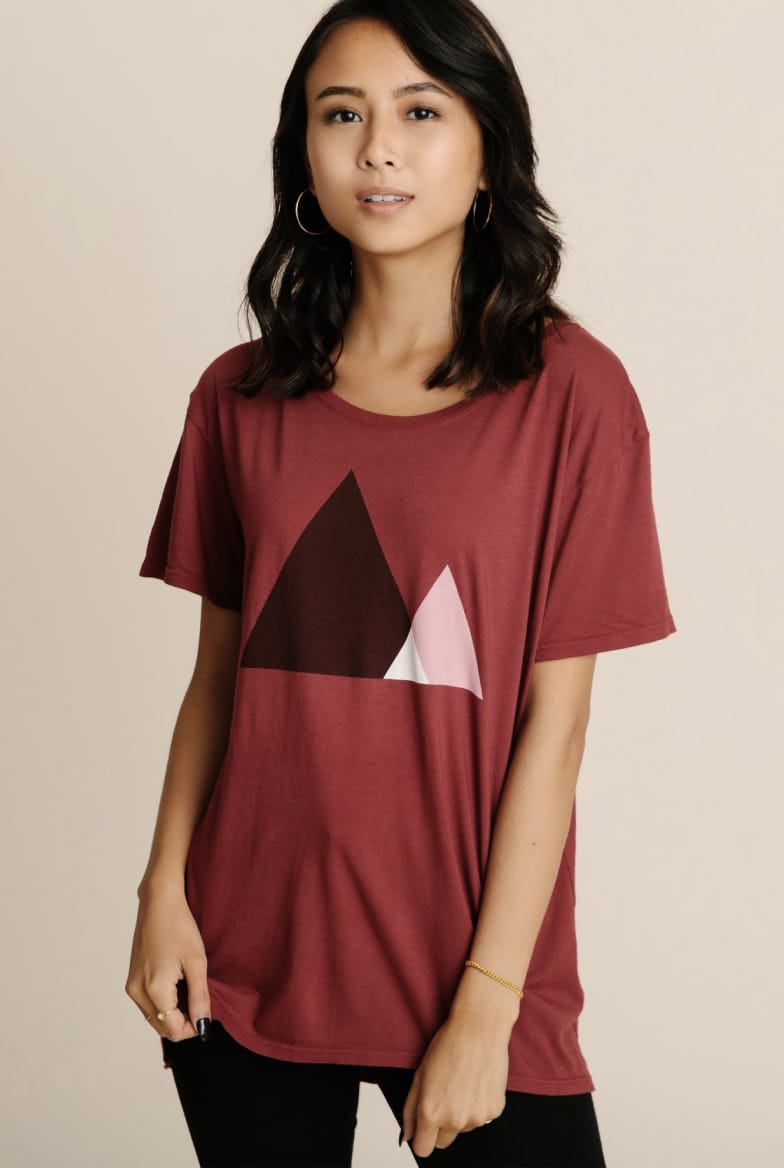 Model wearing women's burgundy red crewneck artwork tee with small white triangle overlapping larger black triangle.